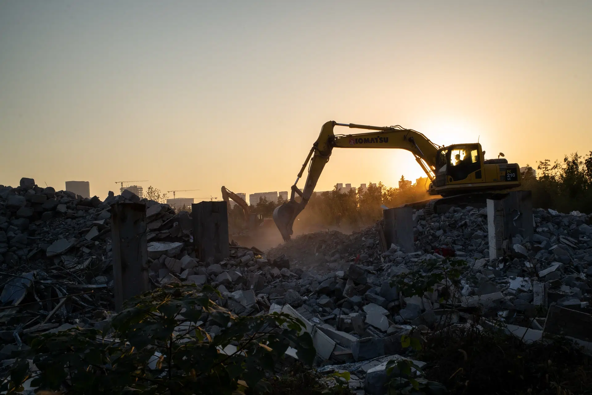 Excavator lucrează pe un teren demolat, la apus, cu orașul în fundal